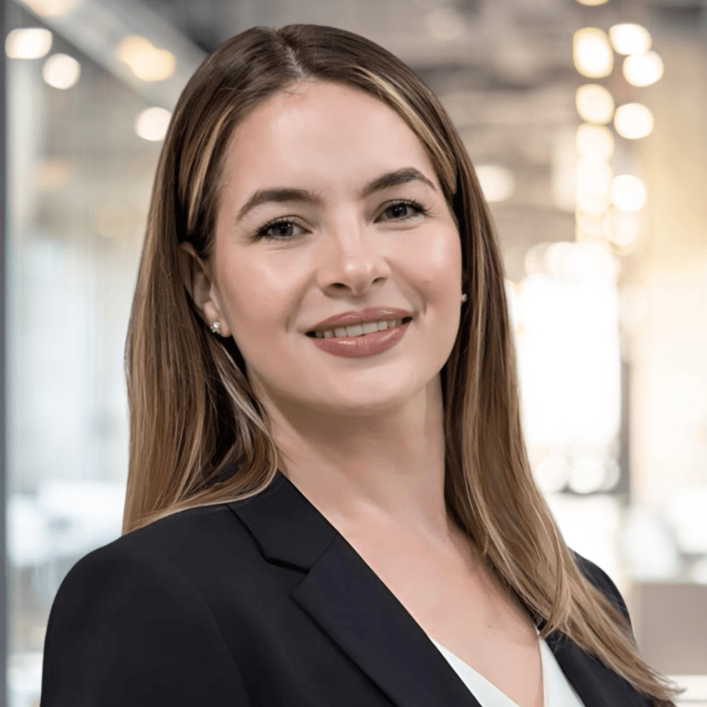 A woman with straight, light brown hair and fair skin smiles while wearing a black blazer and white top, standing in a modern, brightly lit office environment.