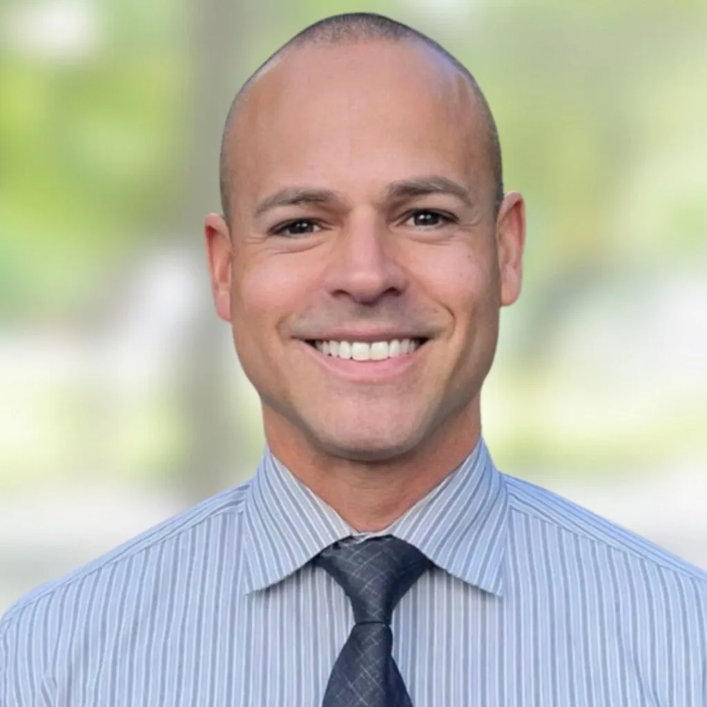 A smiling man with a shaved head wearing a light gray striped shirt and a dark gray tie, standing in front of a blurred green and white outdoor background.