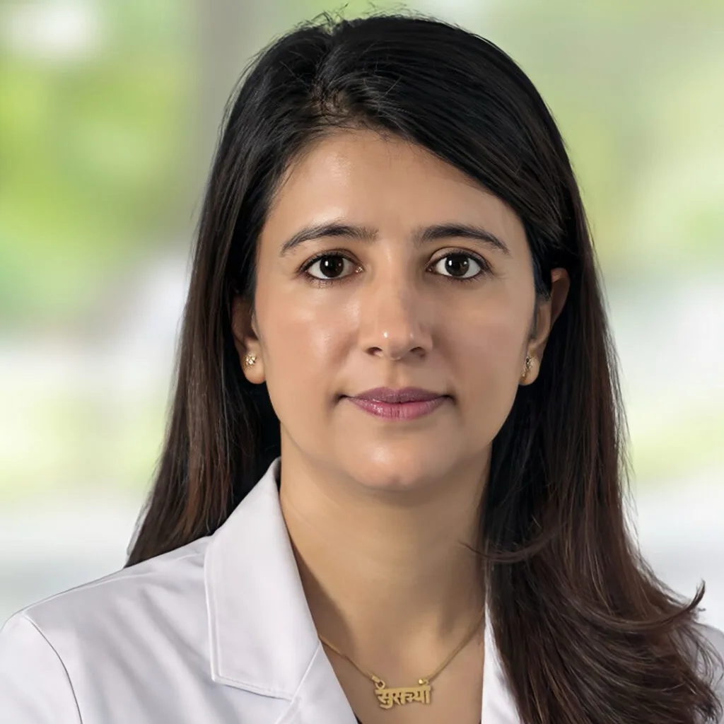 A woman with long dark hair, wearing a white coat and a gold necklace with Hindi script, poses in front of a blurred outdoor background.