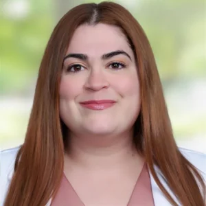 A woman with long straight brown hair, light skin, and a light pink top smiles at the camera against a softly blurred green and white background.