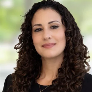 A woman with long, dark, curly hair is smiling softly. She is wearing a black top and a gold necklace, standing in front of a blurred, light-colored background.