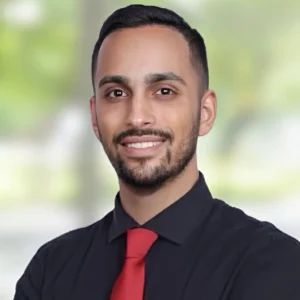 A man with short dark hair, trimmed beard, and mustache smiles at the camera. He is wearing a black shirt and a red tie, with a blurred green and white background.