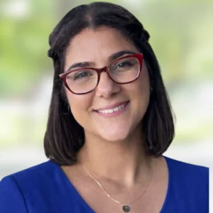 A woman with short dark hair and red glasses smiles at the camera. She is wearing a blue top and a gold necklace, with a blurred green and white background behind her.
