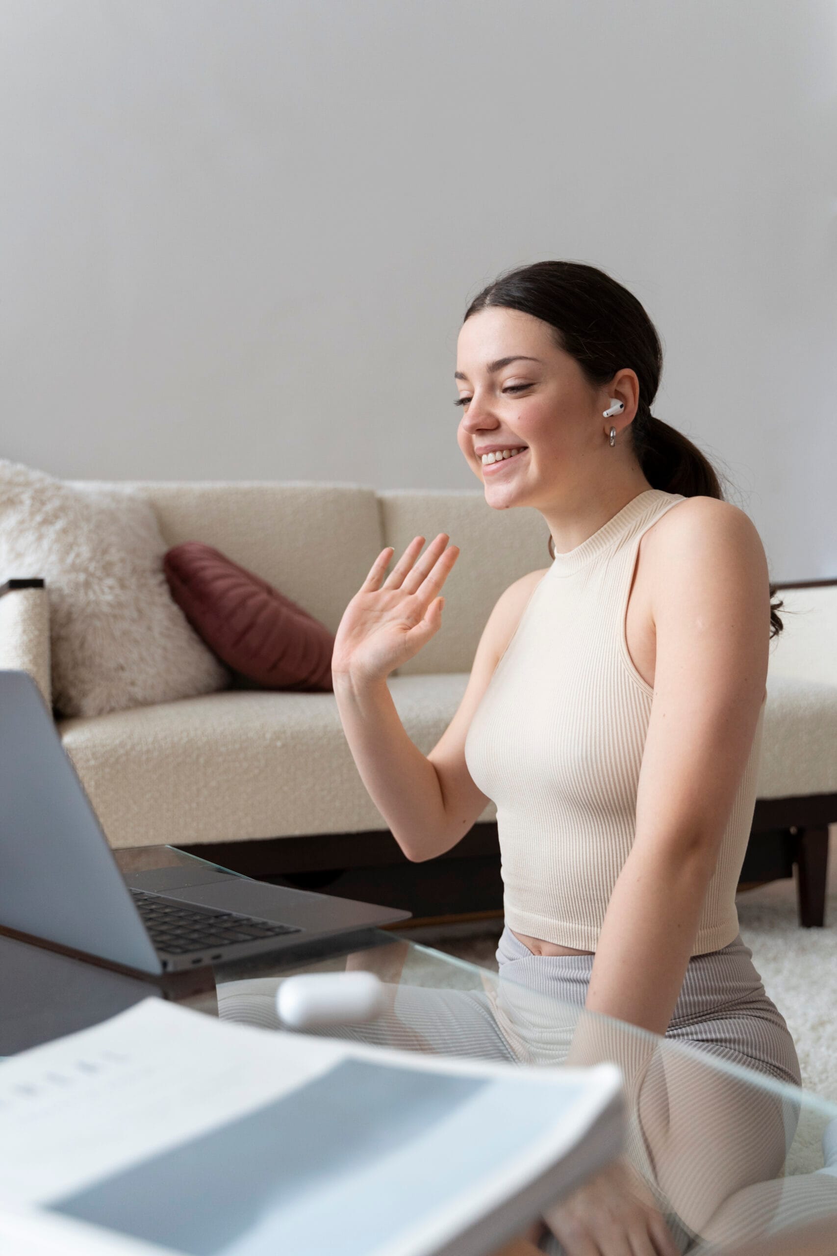 A young woman sits indoors, smiling and waving at her laptop during a video call—perhaps connecting with therapy and psychiatry near me. She wears a sleeveless beige top, her long dark hair tied back, with a sofa and pillow in the background.
