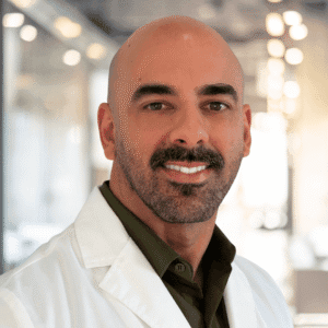 A man with a bald head, trimmed beard, and mustache is wearing a white lab coat and dark shirt, smiling in a bright, modern indoor setting.