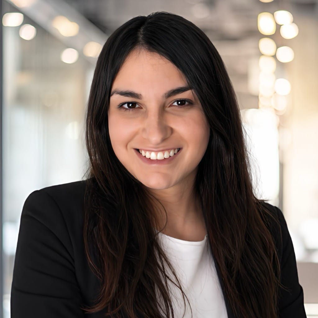 A woman with long dark hair, wearing a black blazer and white top, smiles confidently while standing in a modern, well-lit office hallway with blurred lights in the background.