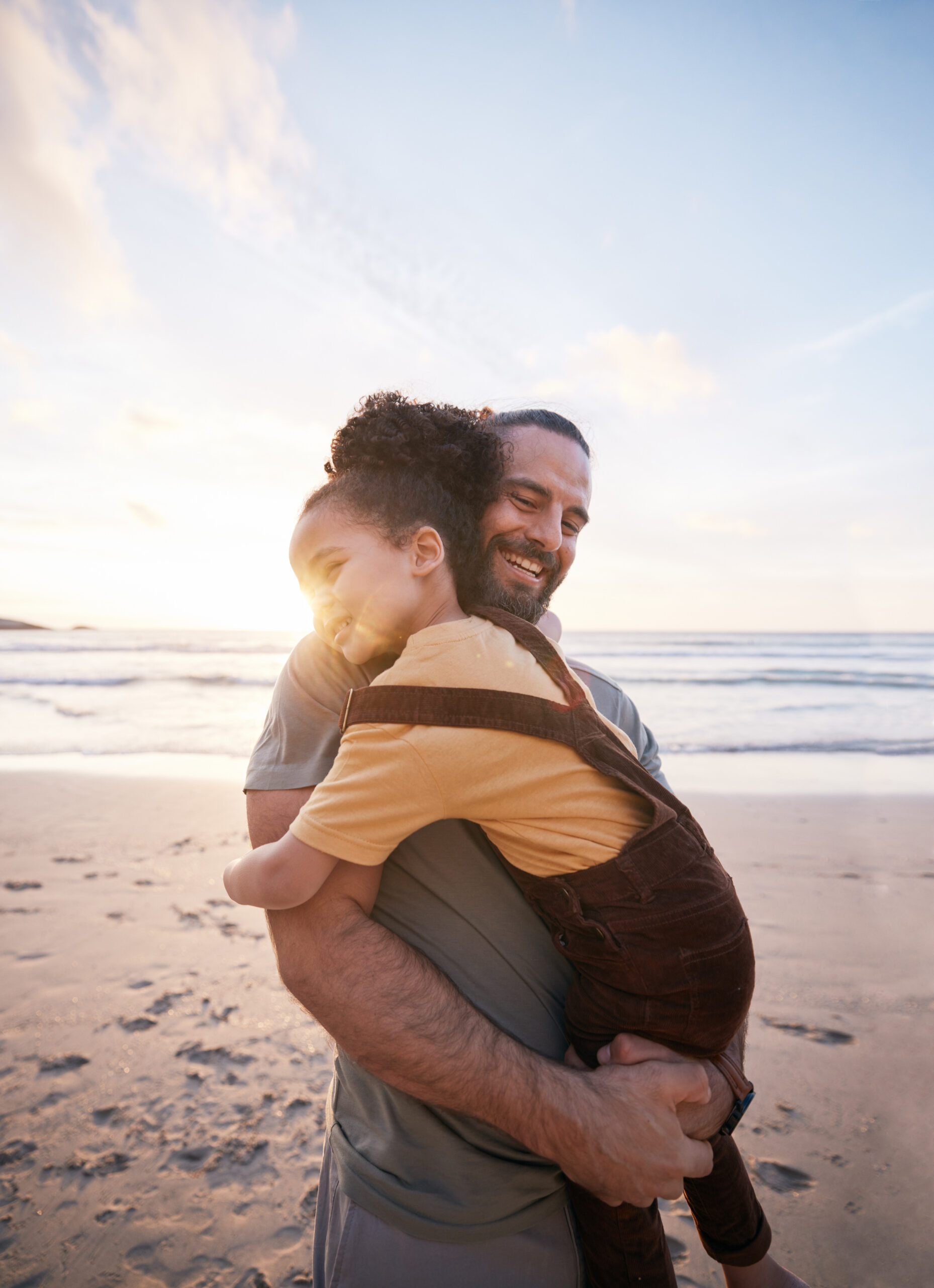 A man smiles as he hugs a child on a sandy beach at sunset. The sun glows behind them, creating a warm and joyful atmosphere by the ocean—capturing moments of peace like those found through therapy and psychiatry near me.